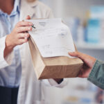 Pharmacist hands prescription medicine to customer in paper bag.