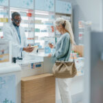 Pharmacy Drugstore Checkout Counter: Professional Black Pharmacist Provides Service, Sells Medicine to Senior Woman Customer. She is Paying with Use of Contactless Payment Credit Card and Part D coverage