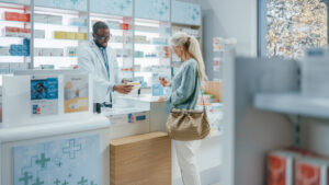 Pharmacy Drugstore Checkout Counter: Professional Black Pharmacist Provides Service, Sells Medicine to Senior Woman Customer. She is Paying with Use of Contactless Payment Credit Card and Part D coverage