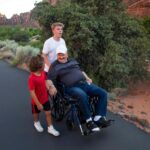 One teenager and one younger boy taking an older, senior man in a wheelchair on a walk on a paved trail in a National Park in the American southwest. Many national parks have accessible features.