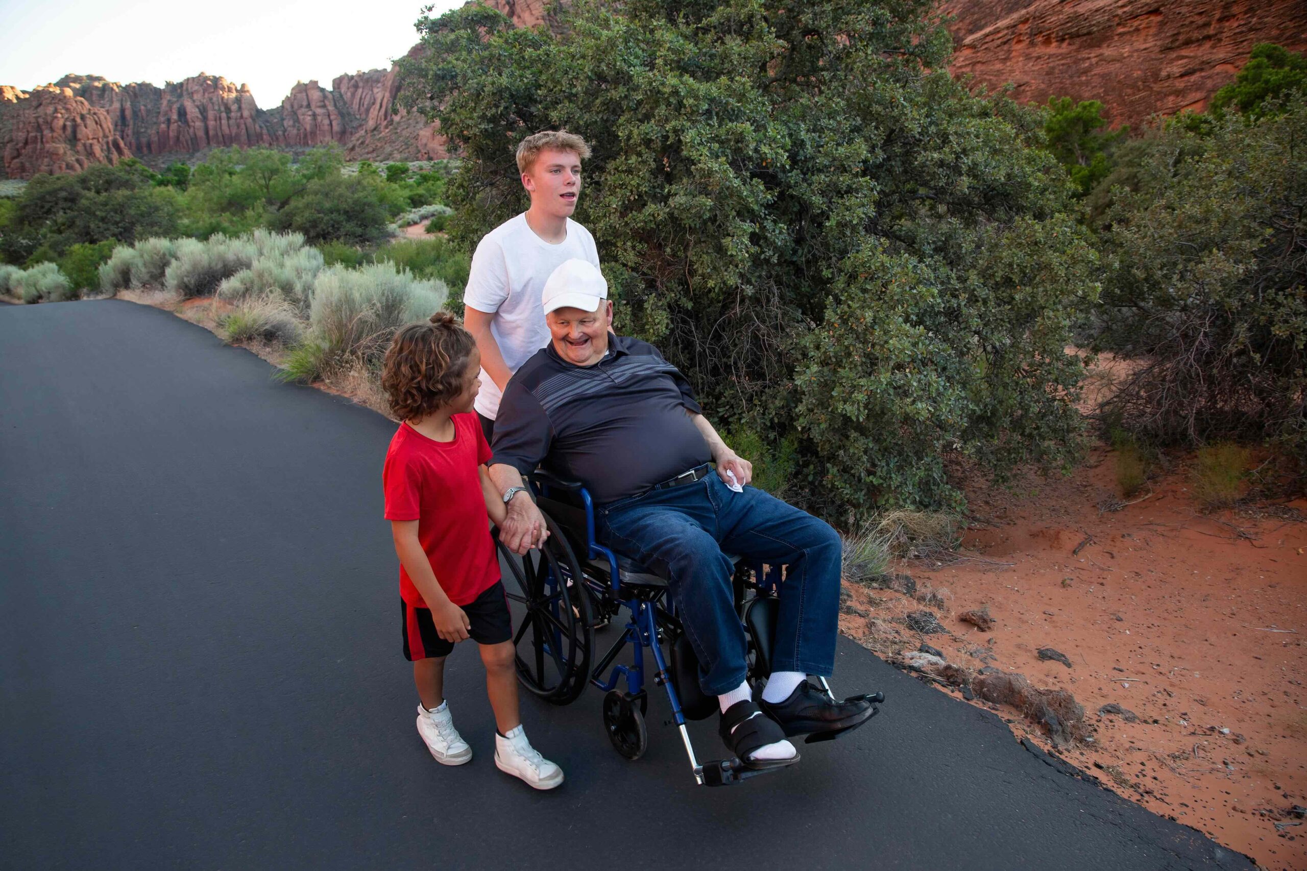 One teenager and one younger boy taking an older, senior man in a wheelchair on a walk on a paved trail in a National Park in the American southwest. Many national parks have accessible features.