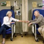 A senior man and woman sitting in chairs holding hands in the hospital, the woman has a canula in her nose, receiving outpatient treatment. When comparing Medicare Supplement Plan G vs. Plan N, the plans cover gaps in coverage for inpatient and outpatient treatment.