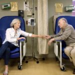 A senior man and woman sitting in chairs holding hands in the hospital, the woman has a canula in her nose, receiving outpatient treatment. When comparing Medicare Supplement Plan G vs. Plan N, the plans cover gaps in coverage for inpatient and outpatient treatment.