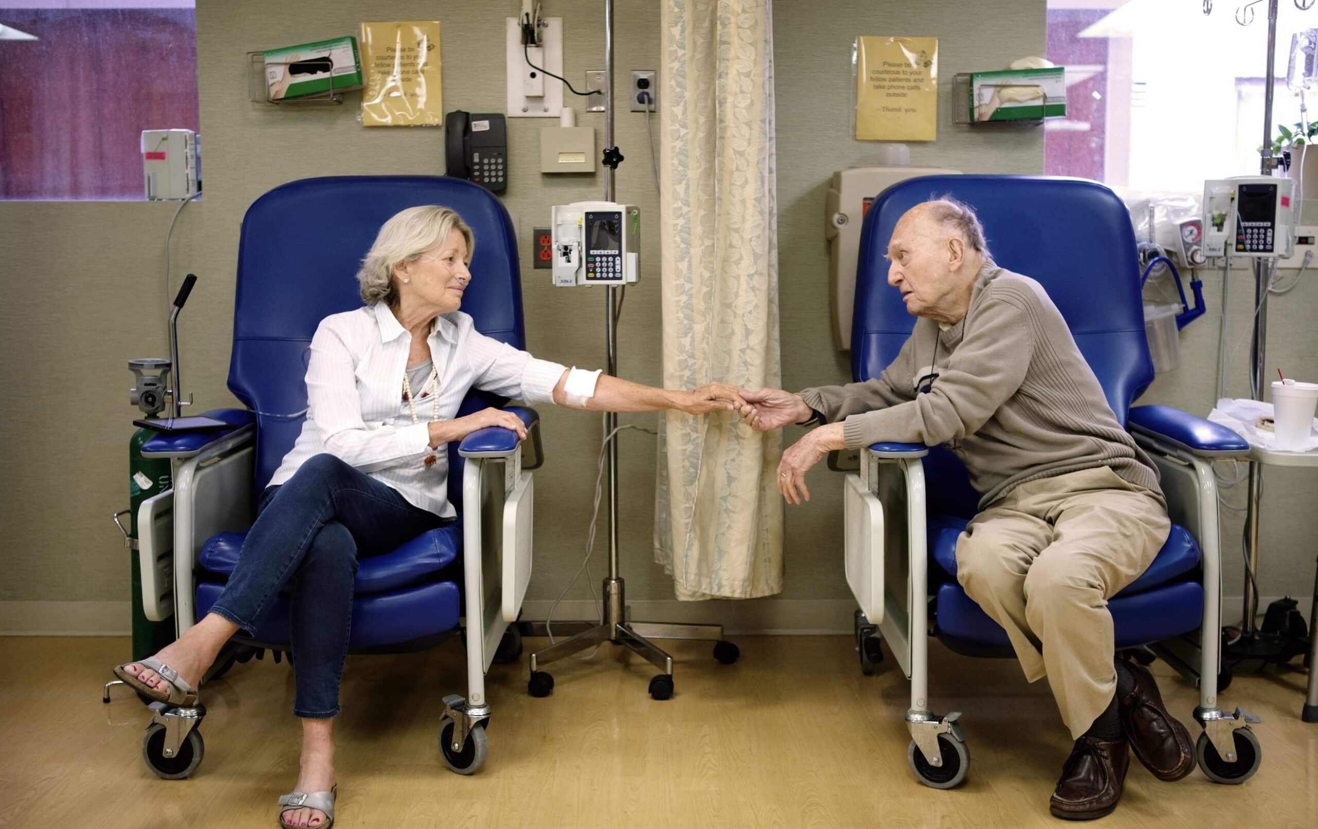 A senior man and woman sitting in chairs holding hands in the hospital, the woman has a canula in her nose, receiving outpatient treatment. When comparing Medicare Supplement Plan G vs. Plan N, the plans cover gaps in coverage for inpatient and outpatient treatment.