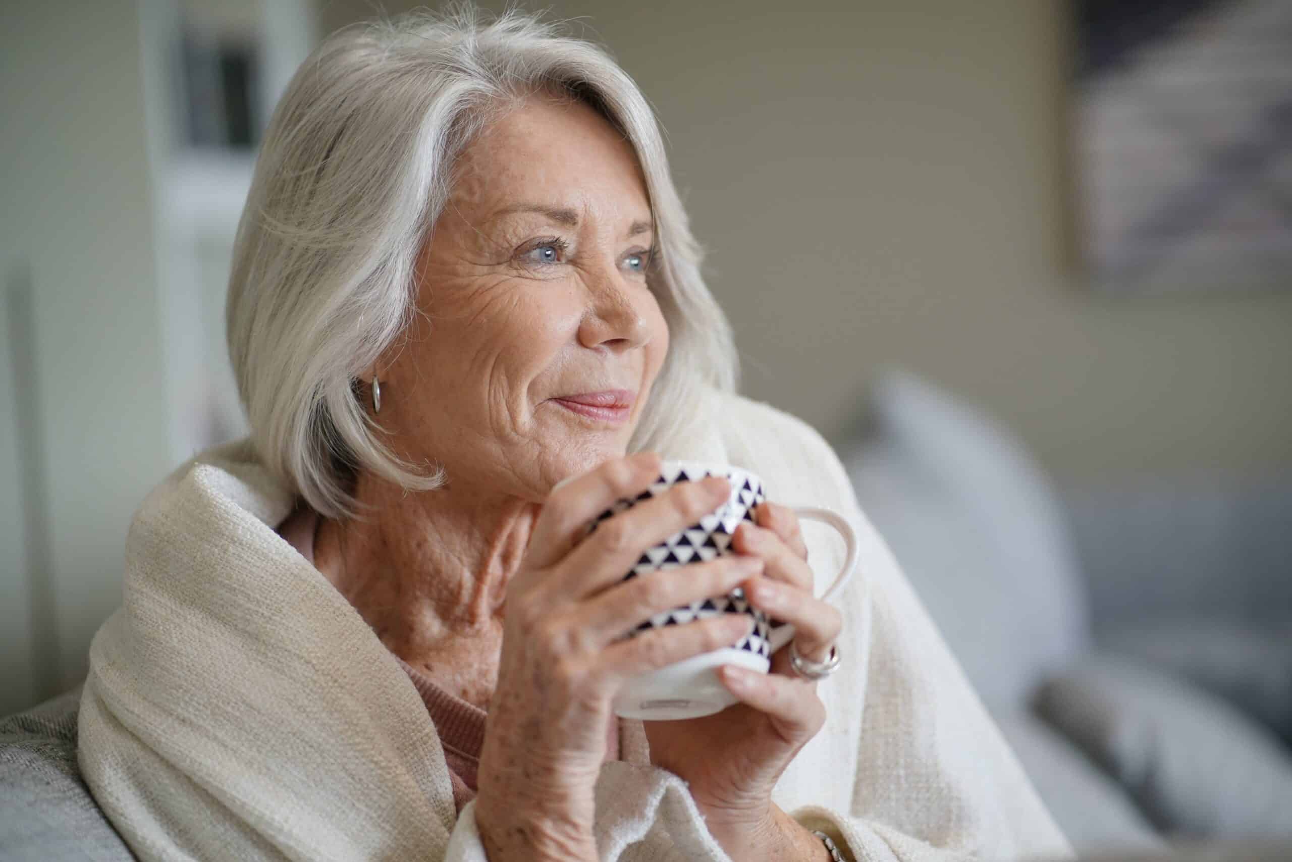Cosy looking senior woman living independently at home — also called aging in place — on the sofa with hot drink
