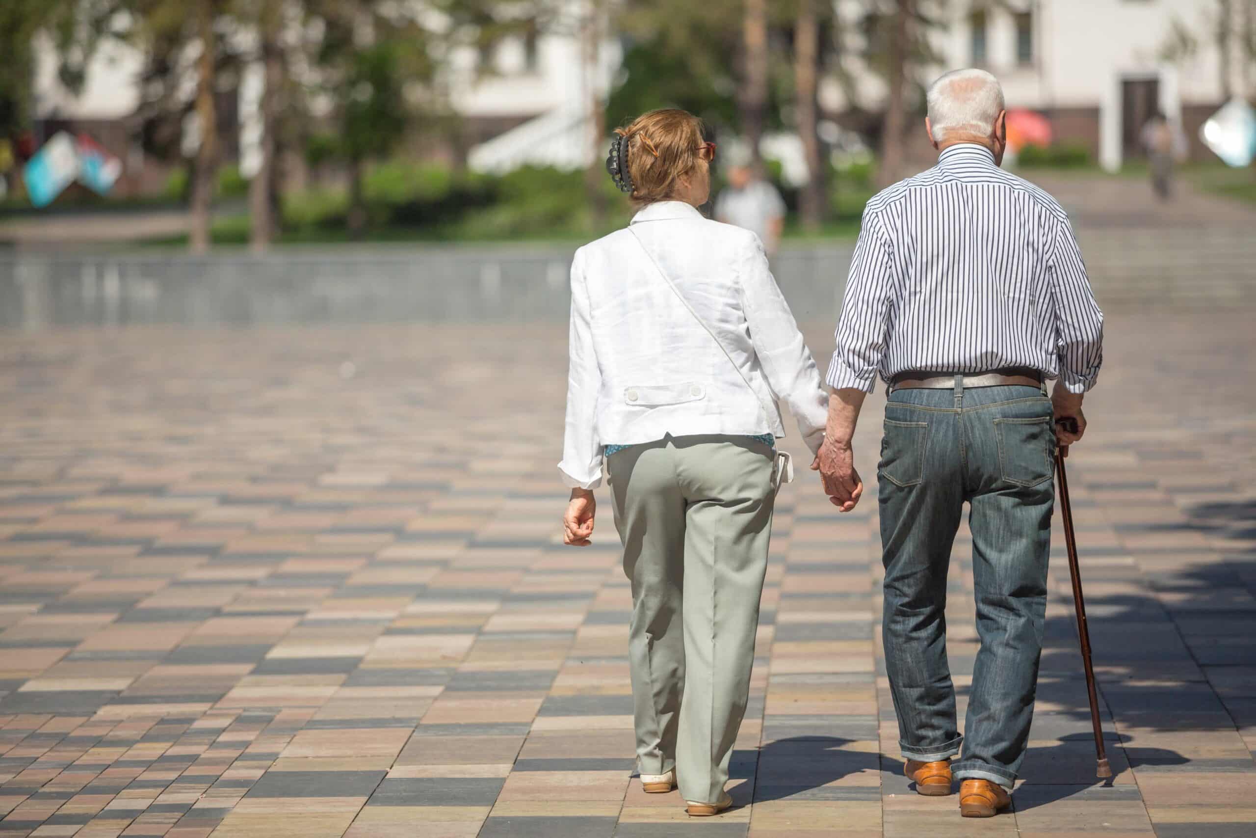 Senior adults walking in a park holding hands. One senior is using a cane.
