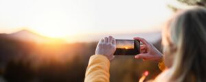 Senior woman hiker standing outdoors in nature at sunset, taking photograph with smartphone.