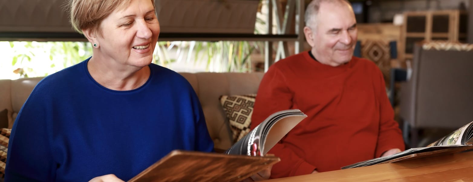 A senior man and woman sit on the same side of a table in a European restaurant, perusing a multi-page menu. Understanding triggers and avoiding them is an important tip for traveling with autoimmune conditions.