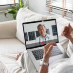 Senior man lounging on sofa with laptop open, video chatting with his physician on a telehealth appointment.