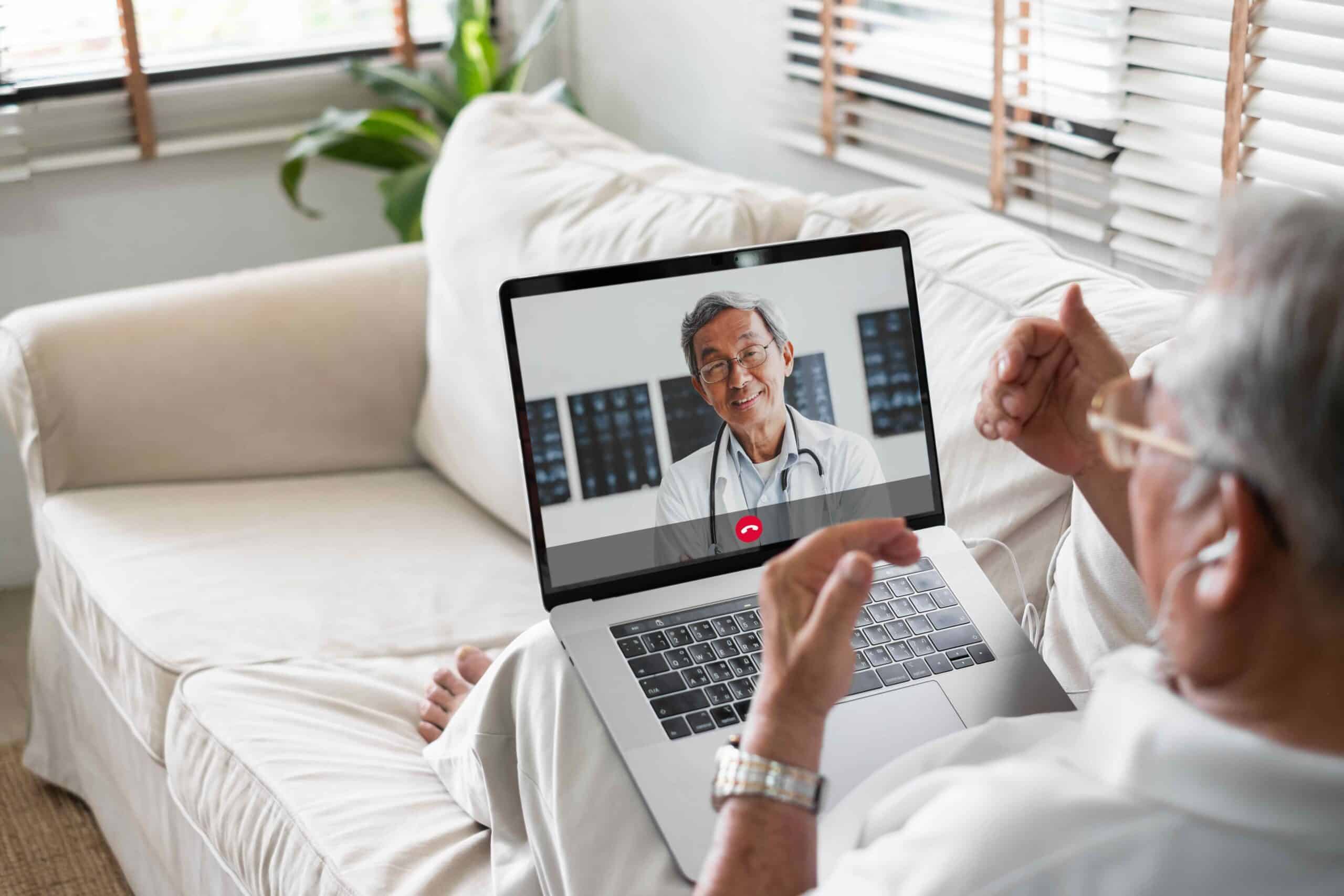 Senior man lounging on sofa with laptop open, video chatting with his physician on a telehealth appointment.