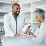 An older woman fills out a form at a pharmacy. A younger pharmacist hands her a paper bag filled with a prescription. Concept for Medicare drug price negotiation program.