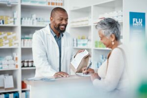 An older woman fills out a form at a pharmacy. A younger pharmacist hands her a paper bag filled with a prescription. Concept for Medicare drug price negotiation program.