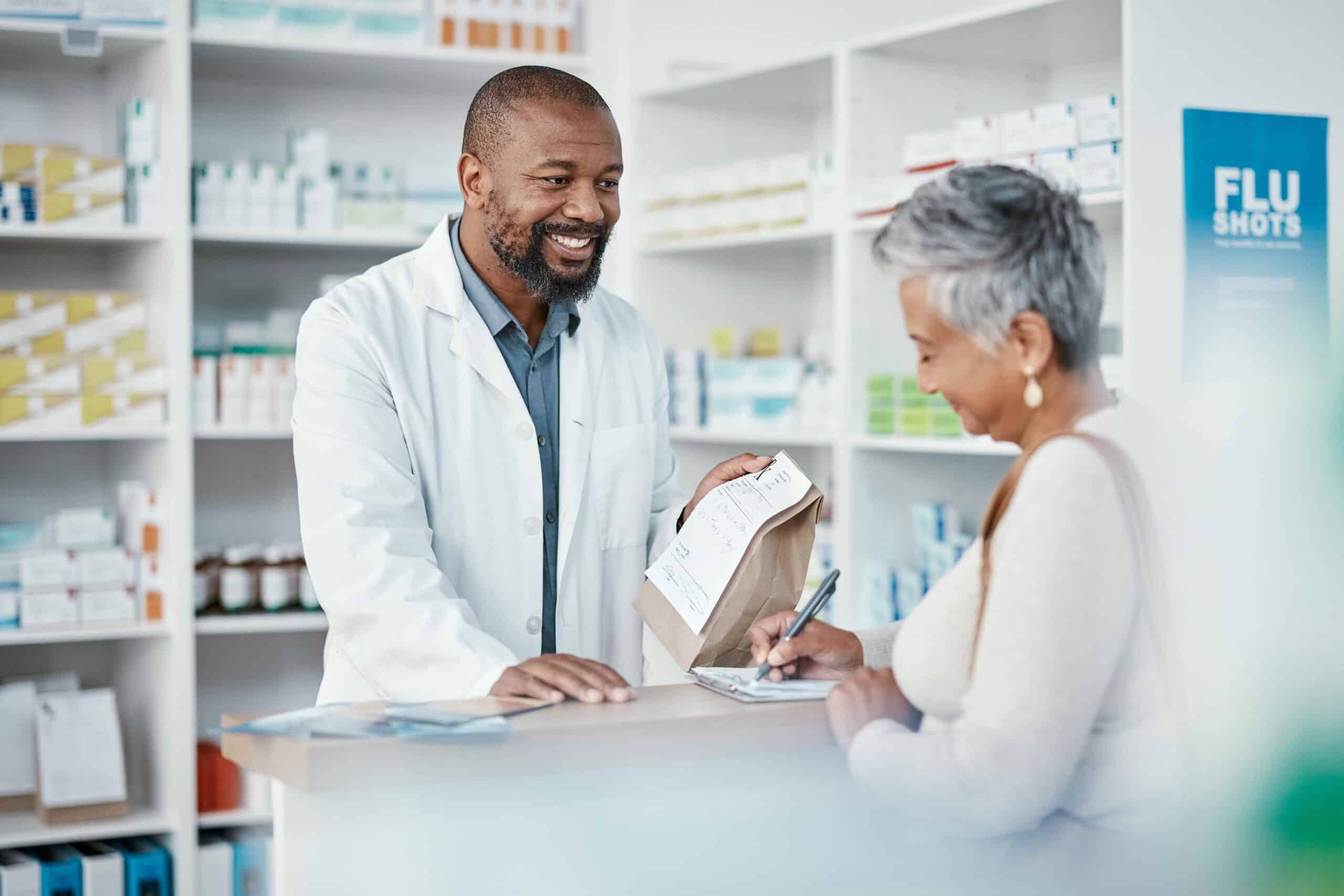 An older woman fills out a form at a pharmacy. A younger pharmacist hands her a paper bag filled with a prescription. Concept for Medicare drug price negotiation program.