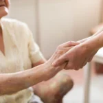 A palliative care specialist holds the hand of a senior patient sitting in a wheelchair.