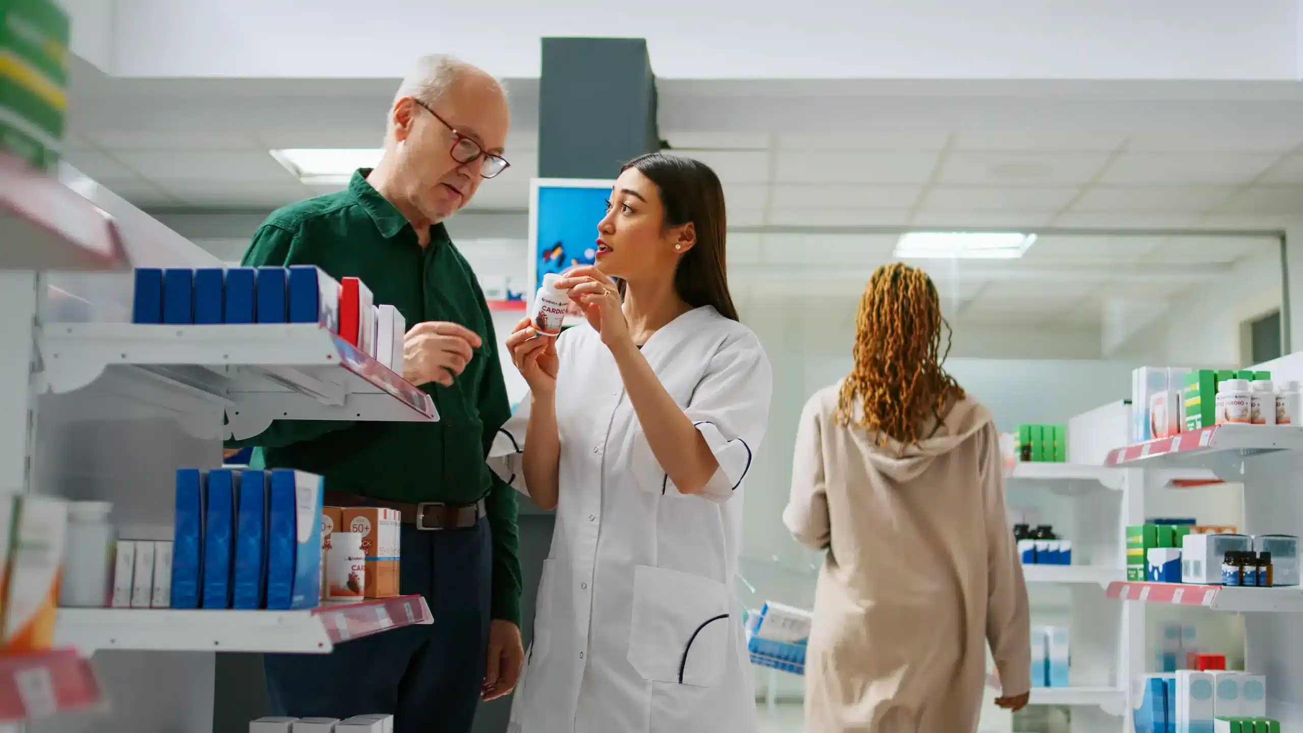 In a brightly lit pharmacy, a female pharmacist in a white lab coat assists an elderly man by discussing a small bottle of medication.