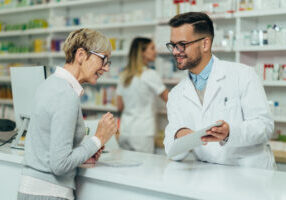 Young male pharmacist giving prescription medications to senior female customer in a pharmacy with female pharmacist in the background