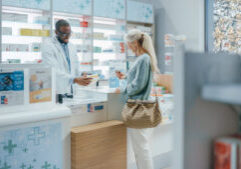 Pharmacy Drugstore Checkout Counter: Professional Black Pharmacist Provides Service, Sells Medicine to Senior Woman Customer. She is Paying with Use of Contactless Payment Credit Card and Part D coverage